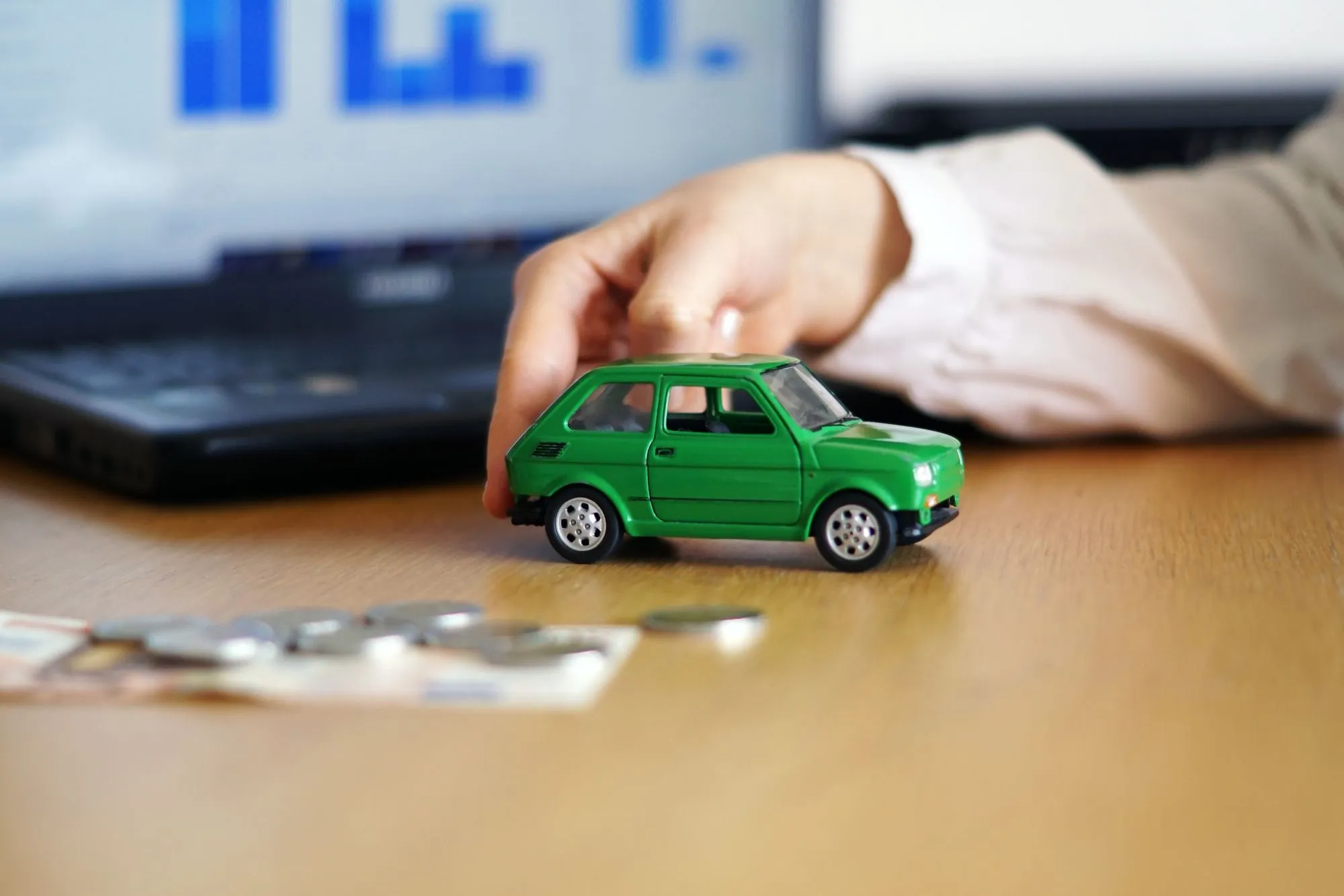 A hand holding a toy car beside scattered coins, symbolizing the cost of insuring different vehicle models